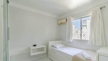 Habitació petita, de color blanc, amb un llit individual, una finestra, una taula de nit i un aparell d'aire condicionat.
