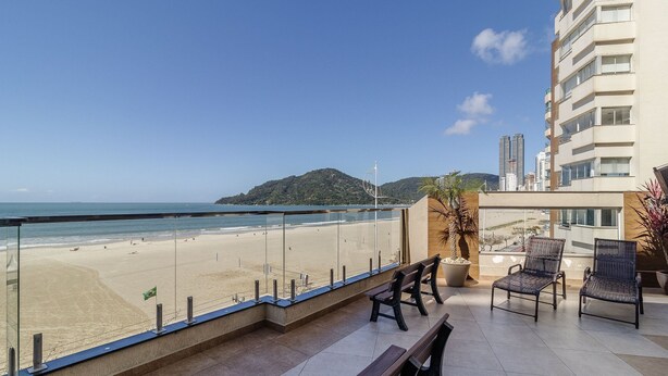 A balcony with a view of a beach and a mountain. There are chairs and a potted plant.