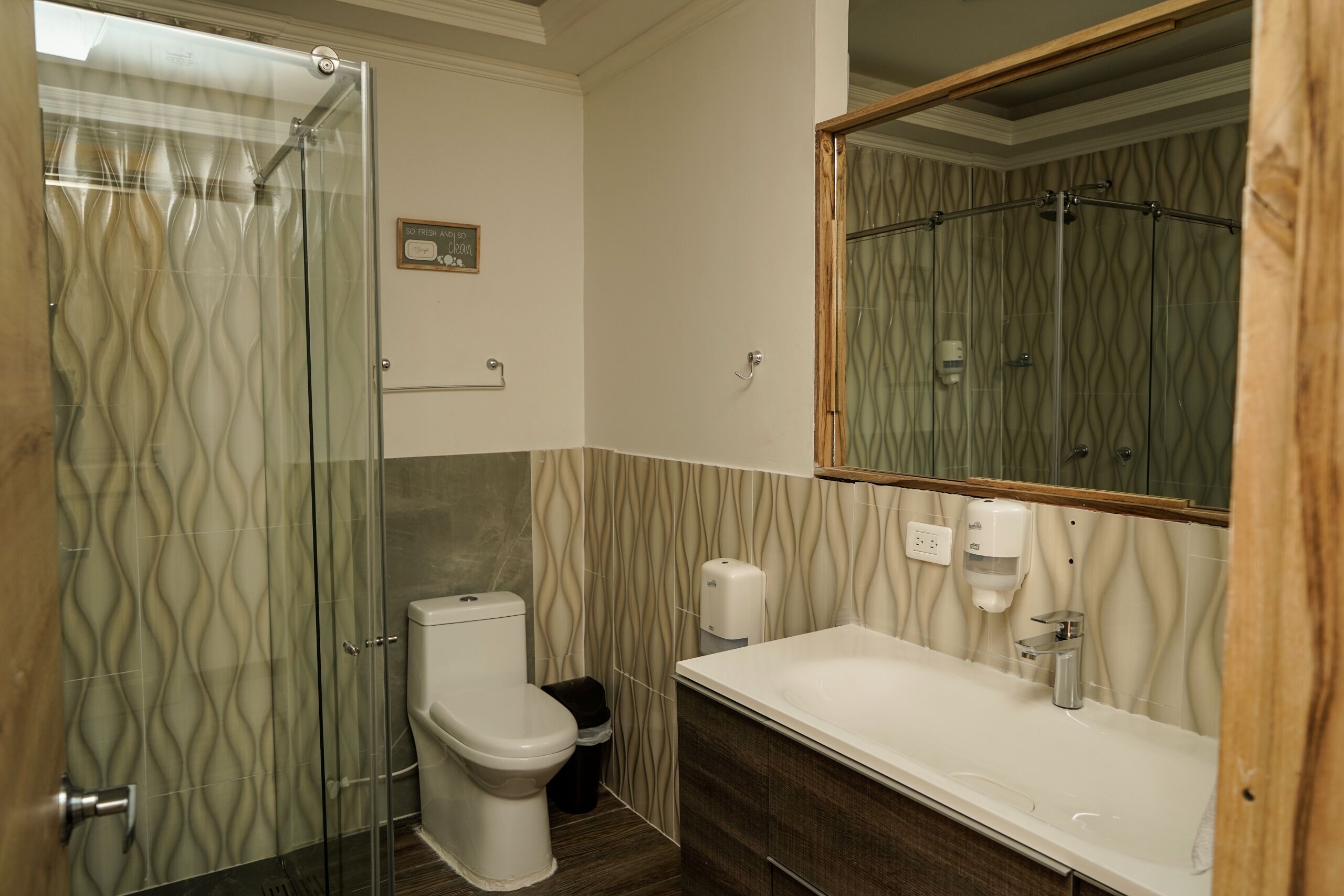 Un baño con cabina de ducha de vidrio, inodoro, lavamanos con dispensador de jabón y espejo.