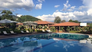 A swimming pool with lounge chairs and umbrellas, surrounded by buildings with red roofs and greenery.