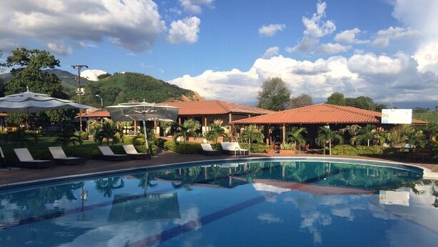 A swimming pool with lounge chairs and umbrellas, surrounded by buildings with red roofs and greenery.