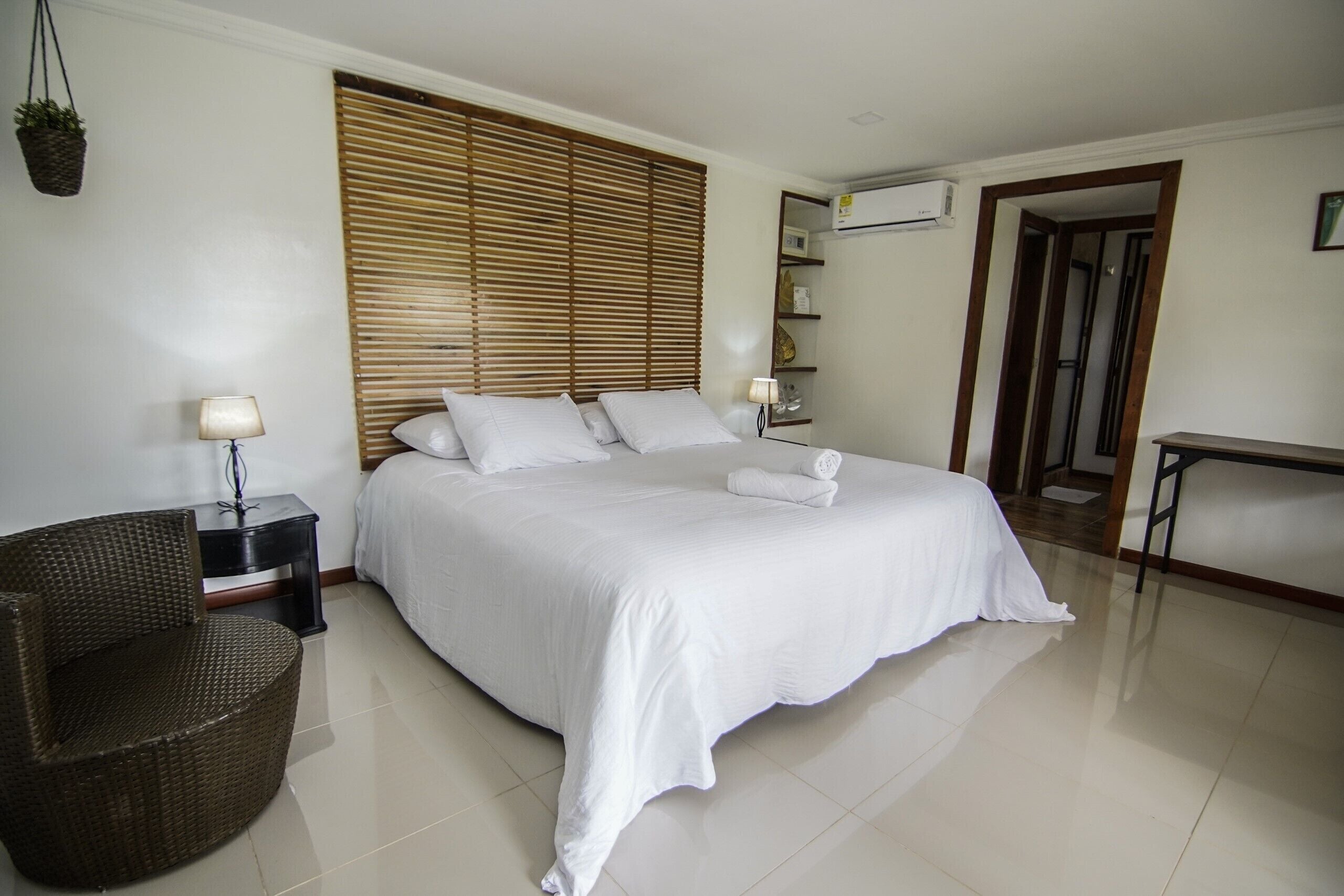 A hotel room with a large bed, a wicker chair, a bedside table with a lamp, and a wooden headboard.