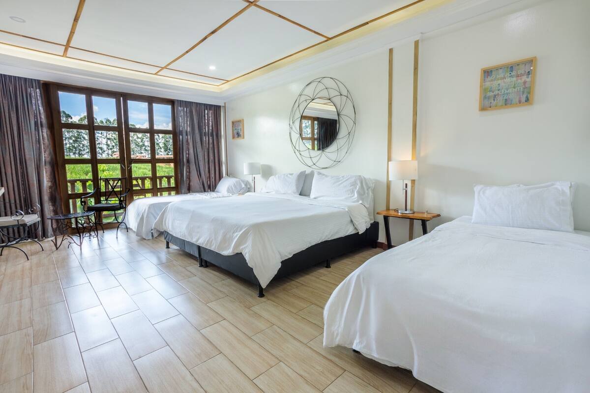 Habitación de hotel con dos camas, un ventanal con cortinas, una mesa de comedor y vistas al exterior con vegetación.