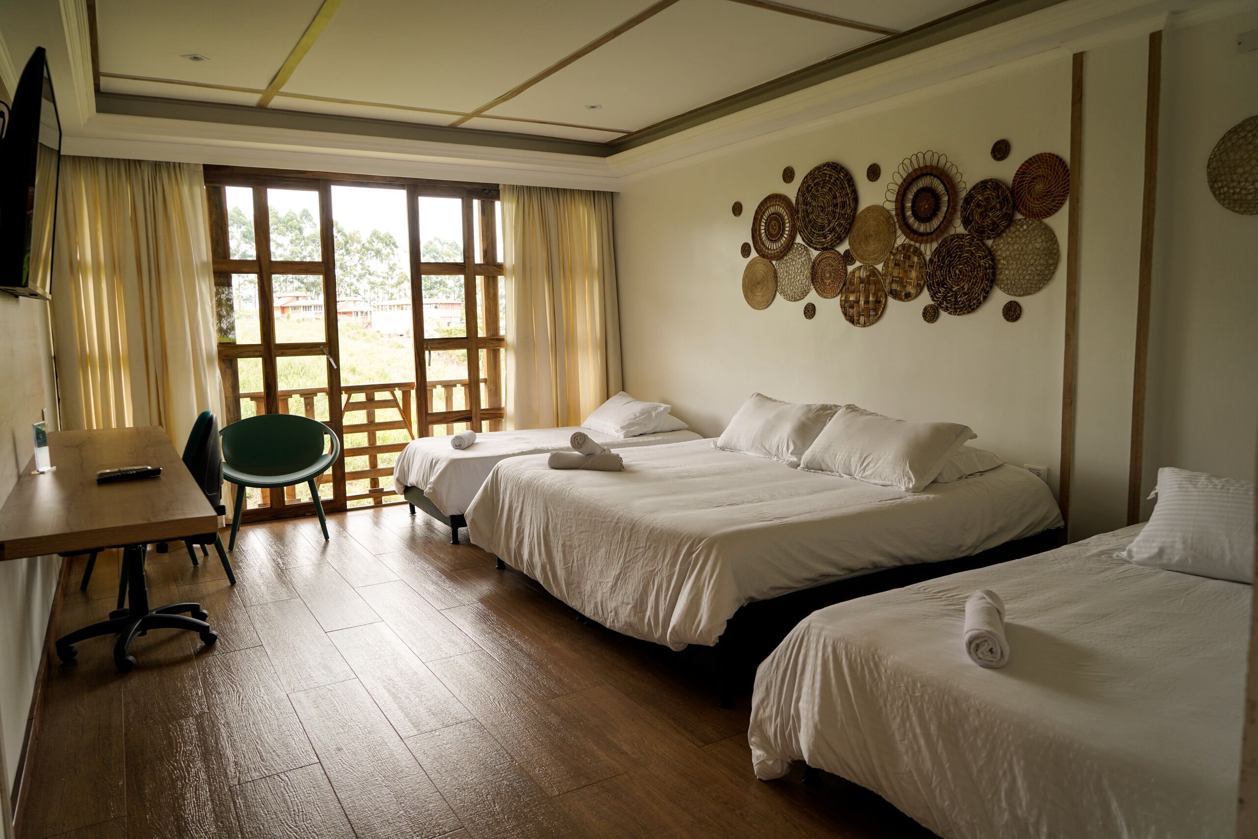 Habitación de hotel con dos camas, un escritorio y una silla. Se aprecia un ventanal con cortinas, y una pared decorada con cestas tejidas.