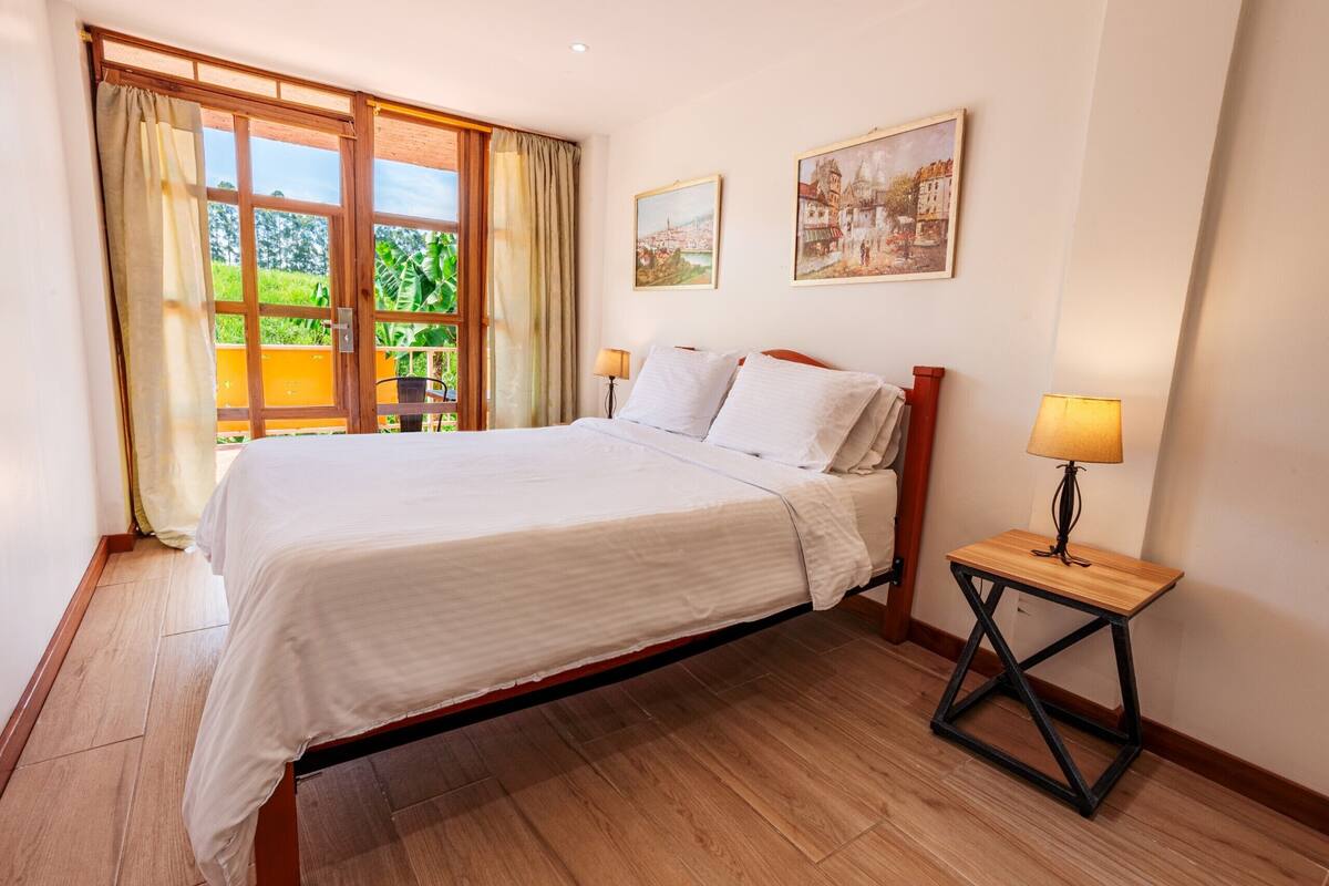 Un dormitorio con una cama grande, suelo de madera, una mesita con una lámpara y dos cuadros en la pared.