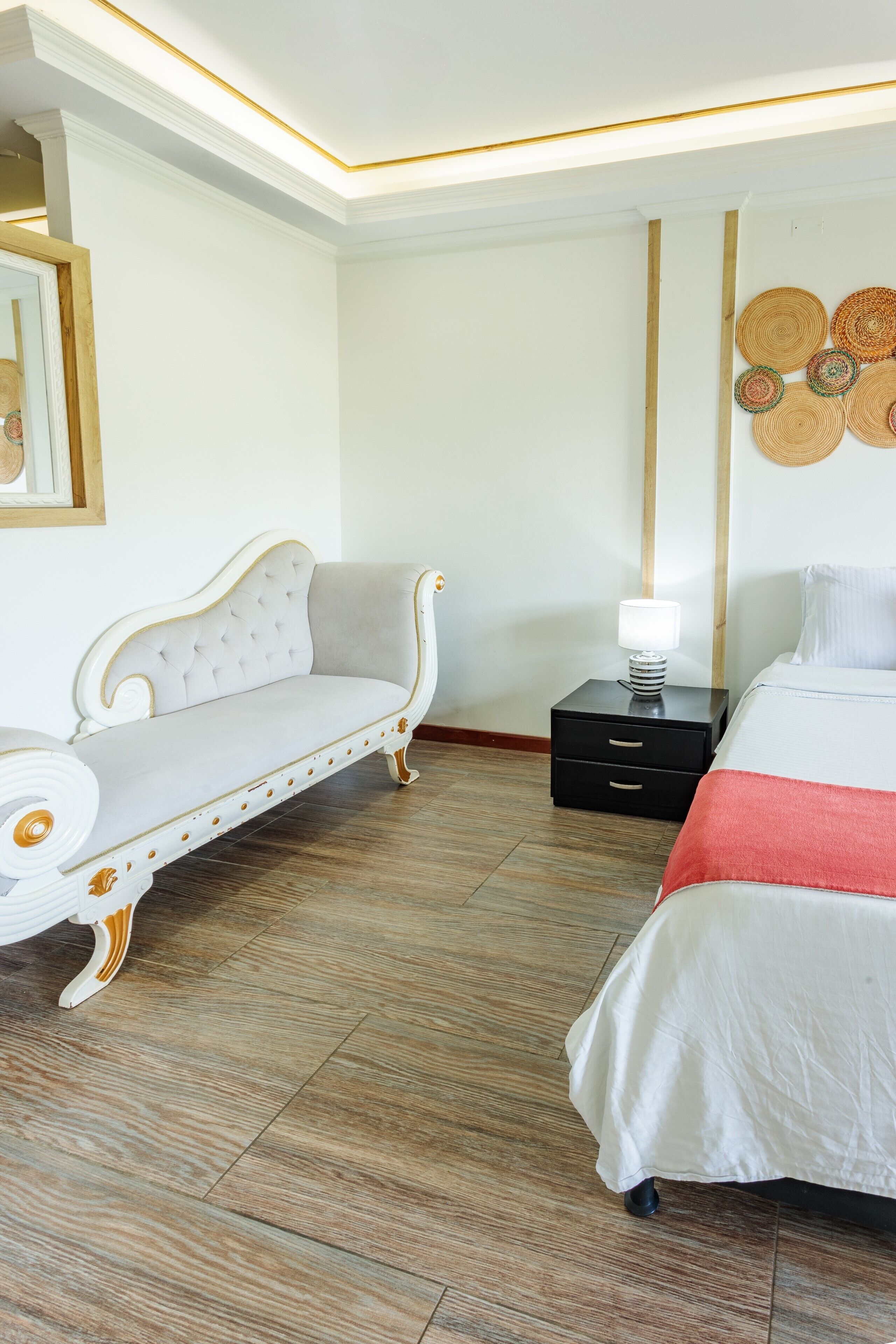 A hotel room with a white sofa, a bed with a red blanket, a nightstand with a lamp, and a wall with woven art.
