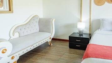 A hotel room with a white sofa, a bed with a red blanket, a nightstand with a lamp, and a wall with woven art.