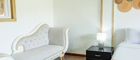 A hotel room with a white sofa, a bed with a red blanket, a nightstand with a lamp, and a wall with woven art.