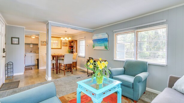 A living room with a blue sofa, a blue armchair, and a glass-top coffee table with a vase of flowers. There is a dining area with a table and chairs visible in the background. - 1 Mi to Sands Beach: Walkable Island Home w/ Yard! (Port Royal)