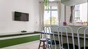 Una cocina moderna con televisor de pantalla plana, gabinetes verdes, sillas blancas y un ventanal con cortinas.