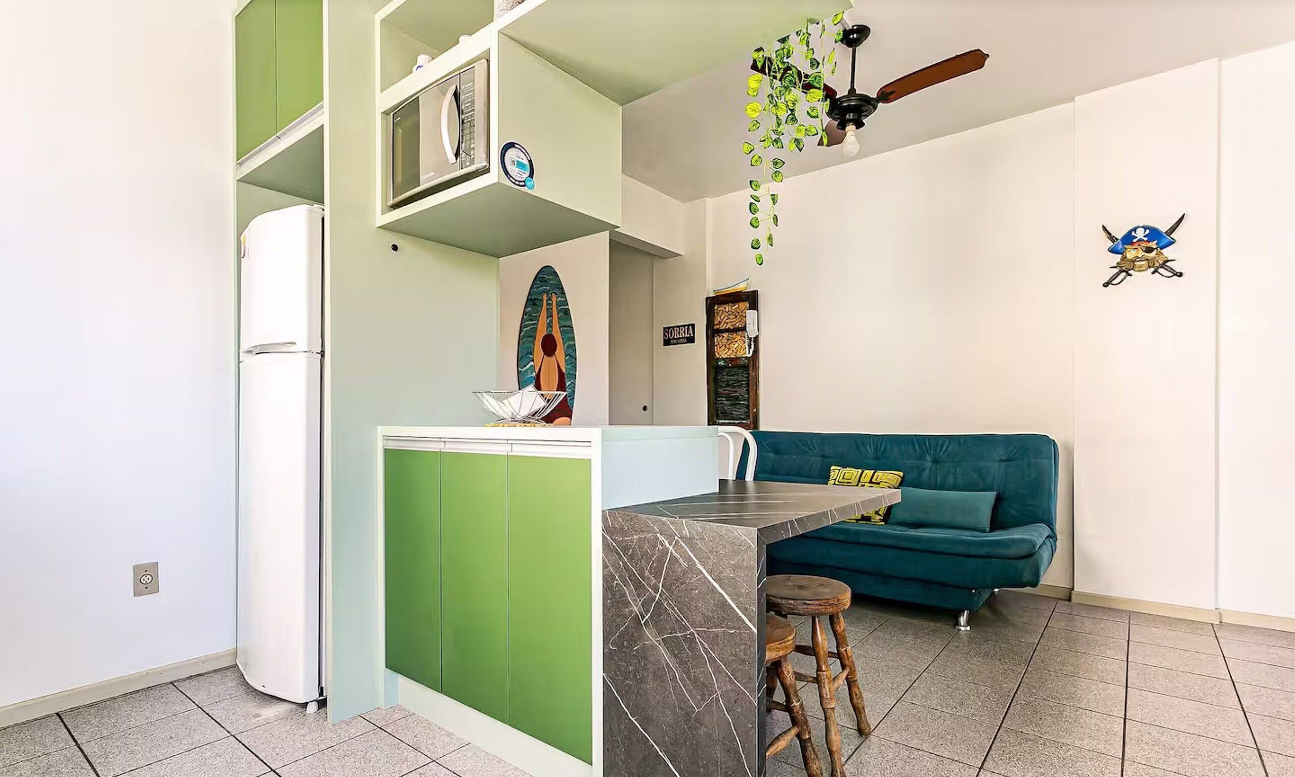 A compact kitchen with a refrigerator, microwave, and a small island with stools. A teal sofa and a ceiling fan are visible in the living area.