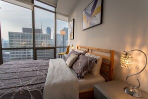 Ein modernes Hotelzimmer mit einem großen Bett, einem Holzkopfteil, einem Nachttisch, einer Lampe und einem Sessel. Ein großes Fenster bietet Blick auf die Stadt.