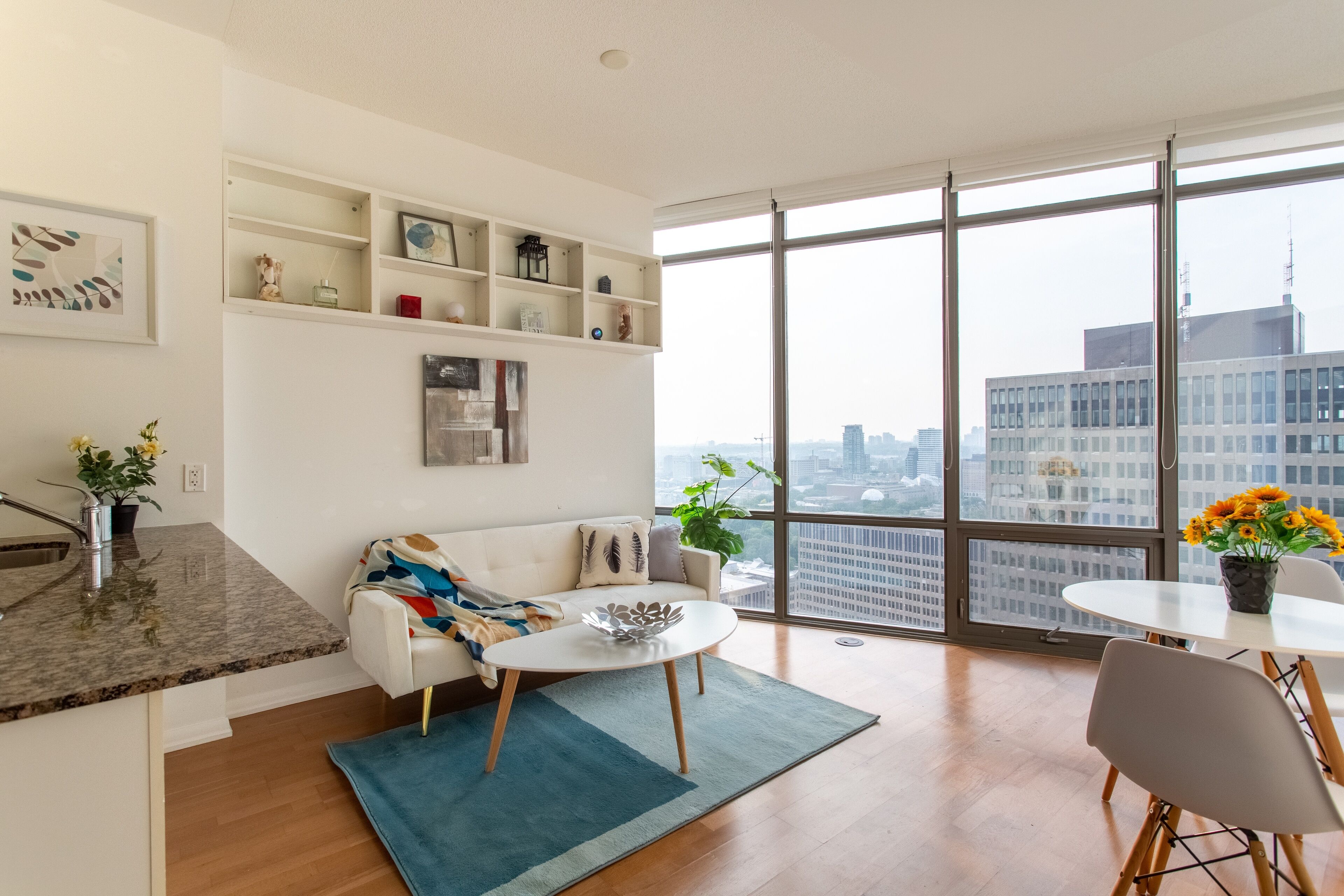 Una sala moderna con un sofá blanco, una mesa de centro y una mesa de comedor con sillas. Hay un ventanal con vistas a edificios y un paisaje urbano.