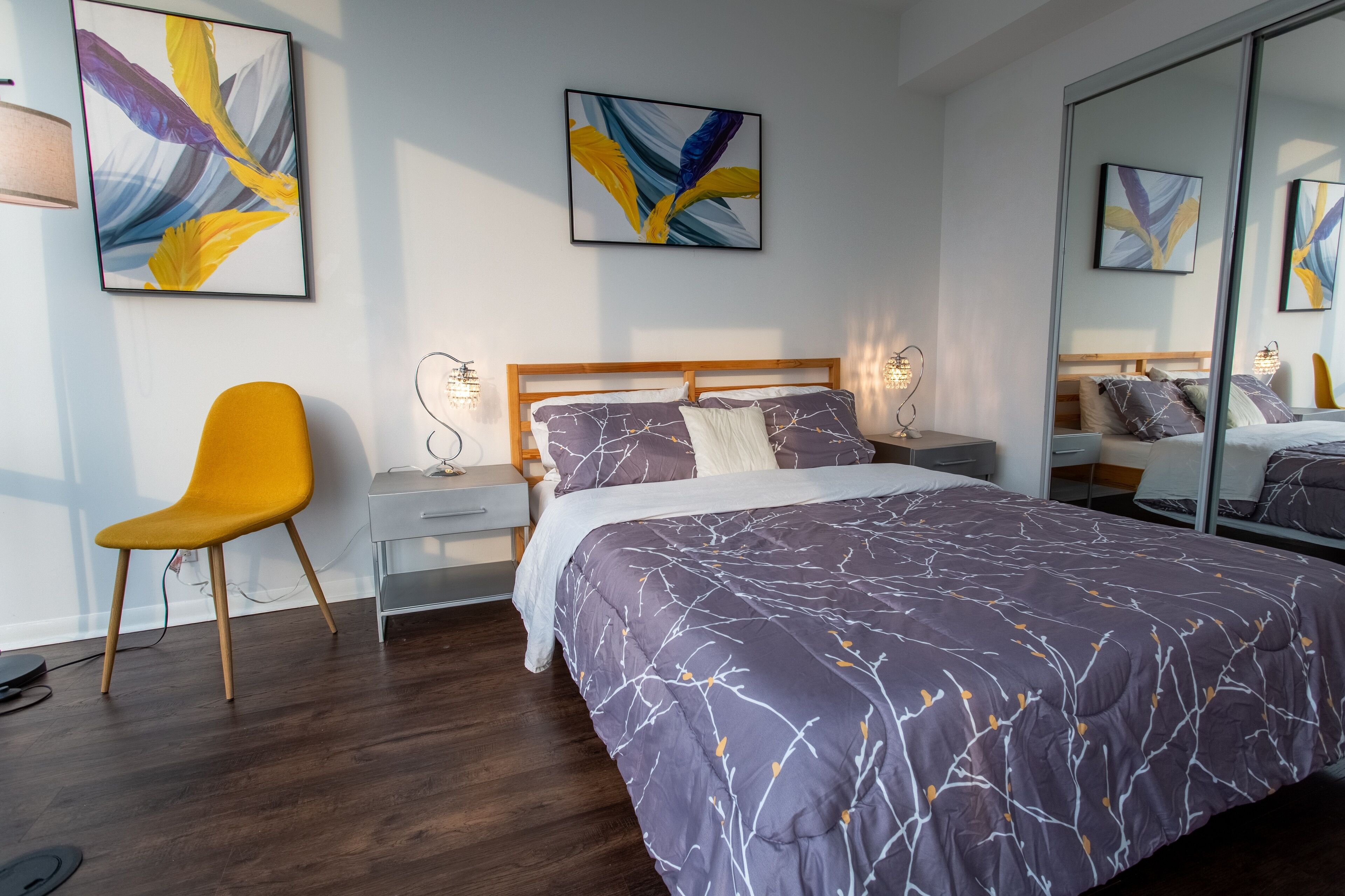 Un dormitorio moderno con cama de cabecera de madera, una mesita de noche con lámpara, una silla amarilla y un cuadro abstracto en la pared.