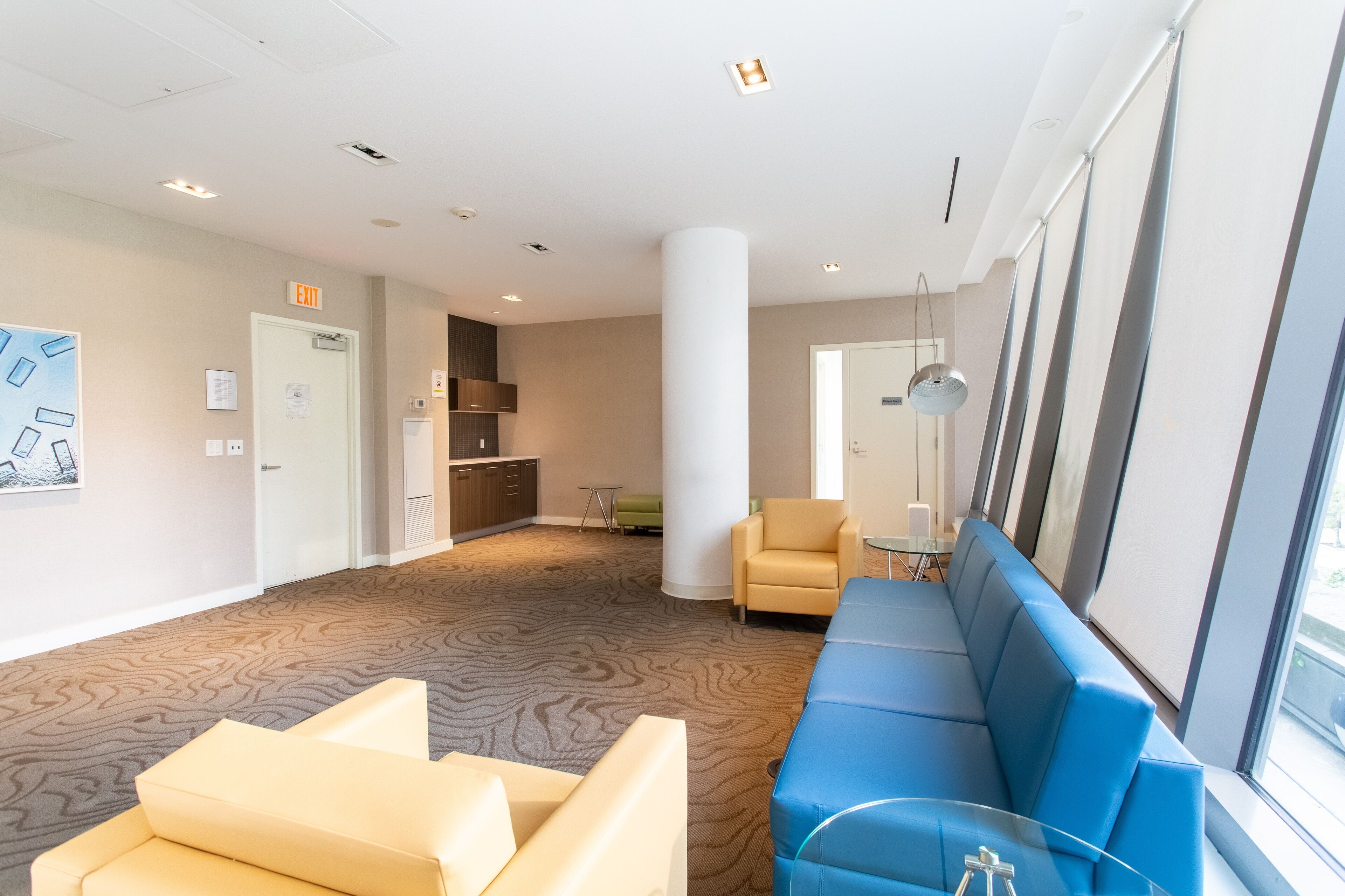 A modern hotel lobby with a patterned carpet, a blue sofa, a beige sofa, a small table, and a kitchen area in the background.