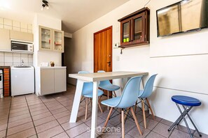 Cocina compacta con microondas, refrigerador y armarios. Hay una mesa de comedor con cuatro sillas y un taburete azul. Se ve una puerta de madera y una ventana pequeña.