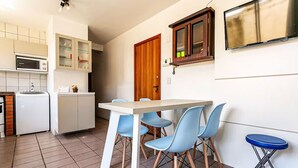 A compact kitchen with a microwave, refrigerator, and cabinets. There is a dining table with four chairs and a blue stool. A wooden door and a small window are visible. - Apart With Barbecue 750 m From the Beach Bou201 (Florianópolis)