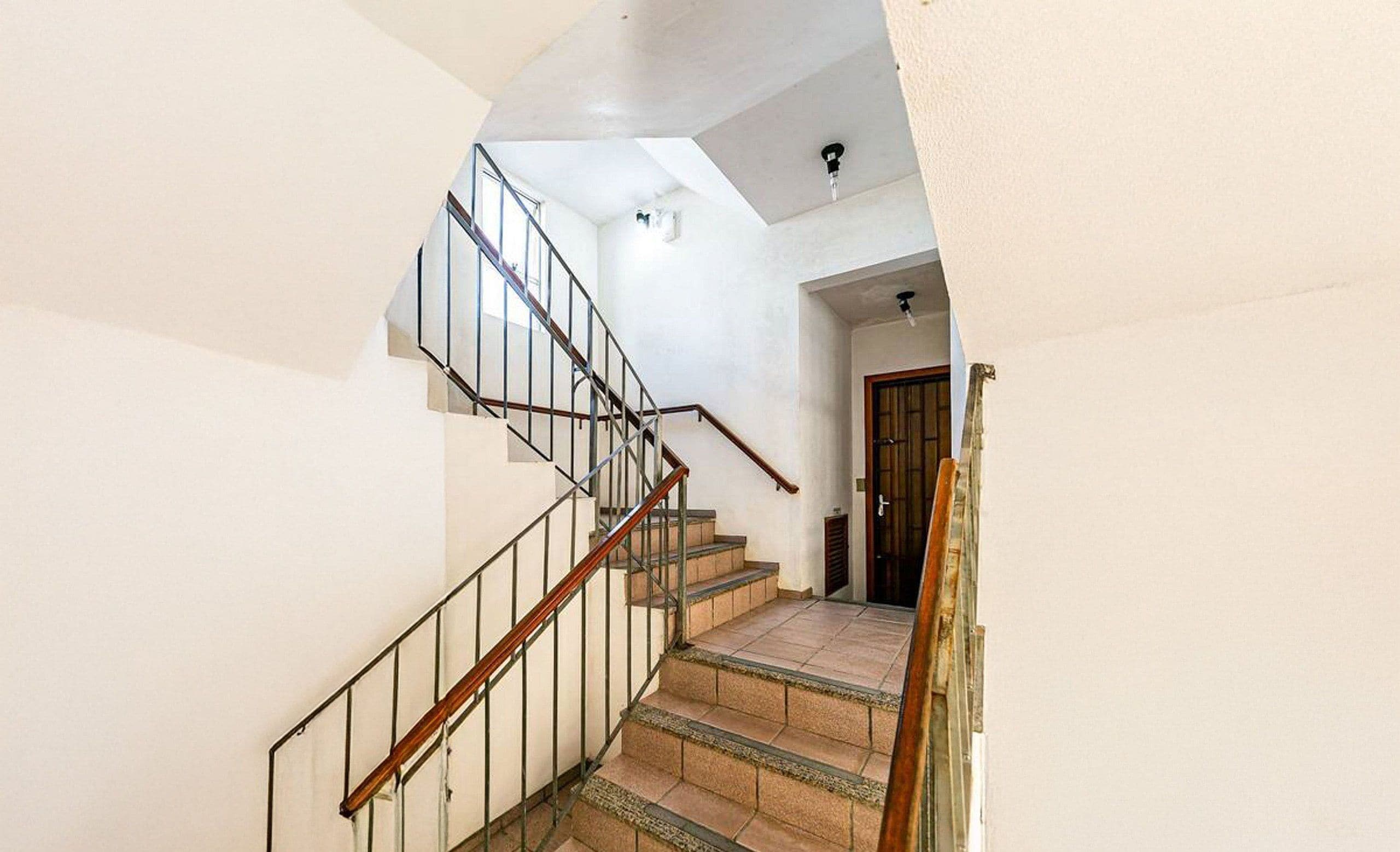 Un escalier avec une main courante en bois et des balustres en métal, menant à une pièce dotée d’une porte en bois et d’un luminaire au plafond.