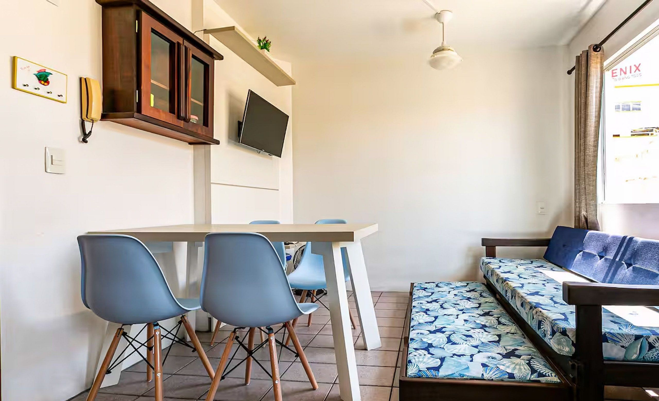 A compact living space with a dining area, a sofa bed with a floral cover, a television mounted above the sofa, and a small shelf with a plant.