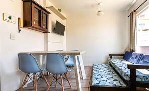 A compact living space with a dining area, a sofa bed with a floral cover, a television mounted above the sofa, and a small shelf with a plant. - Apart With Barbecue 750 m From the Beach Bou201 (Florianópolis)