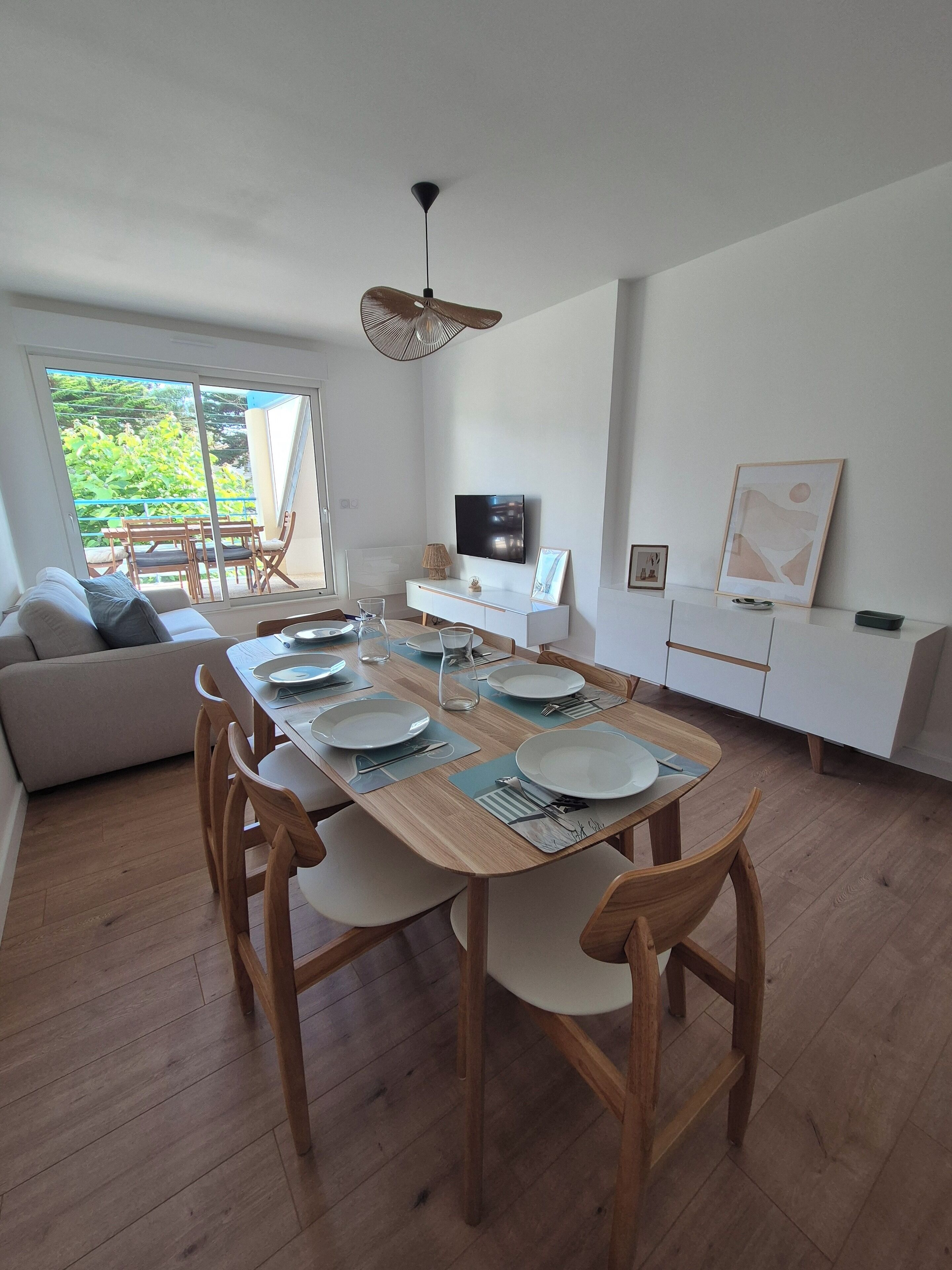 Un espace repas moderne avec une table en bois dressée pour quatre personnes, un canapé et un téléviseur posé sur un meuble blanc.
