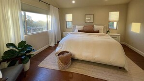 Une chambre avec un grand lit, deux tables de chevet, une fenêtre avec des rideaux, une plante en pot et un panier en osier.