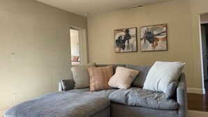 Un canapé d’angle gris, agrémenté de plusieurs coussins, se trouve dans un salon aux murs de couleur beige, où sont accrochées deux peintures abstraites.