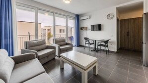 Een moderne woonkamer met een zithoek, een eettafel met stoelen, een aan de muur bevestigde televisie en een groot raam met gordijnen.