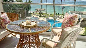 A balcony with a glass table, wicker chairs, and a view of the ocean and palm trees. - Caribbean Sunscape Beachfront Apartment (Playa Juan Dolio)