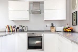 A modern kitchen with white cabinets, a built-in oven, a kettle, and a fruit bowl on the counter. - Contemporary 2-Bed 2-Bath Home with Parking (Gloucestershire)