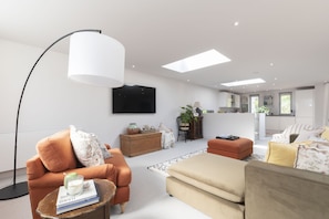 Un salon moderne avec un canapé, un pouf et une table basse, un téléviseur à écran plat, et un coin cuisine visible à travers une porte ouverte.