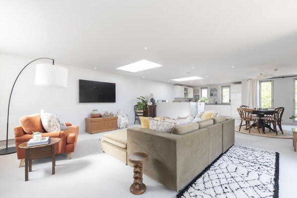 A modern living room with a beige sofa, a round coffee table, and a floor lamp. There is a kitchen area with a dining table and chairs visible in the background. - Contemporary 2-Bed 2-Bath Home with Parking (Gloucestershire)