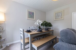 A dining area with a wooden table set for four, surrounded by chairs. There are framed artworks on the walls, a potted plant on the table, and a bench to the right.