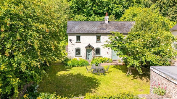 Une maison en pierre avec une porte verte, entourée d’une végétation luxuriante et d’une petite pelouse agrémentée d’une table et de chaises.