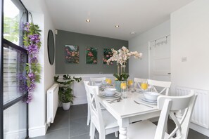 A dining area with a white table set for four, surrounded by white chairs. There are three framed floral paintings on the wall above the table.
