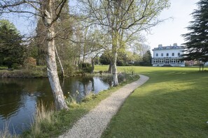 Un sentier paisible le long de la rivière mène à un grand bâtiment blanc à la façade symétrique et à l’entrée centrale.