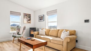 A living room with a beige sofa, a wooden coffee table, a fireplace, and a view of the mountains through large windows. - Cholla Hills Home: Stunning 360° Views, Near JT (Yucca Valley)