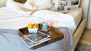 A neatly made bed with white linens, a bedside lamp, a tray with fruits and a mug, and a newspaper. - Cholla Hills Home: Stunning 360° Views, Near JT (Yucca Valley)