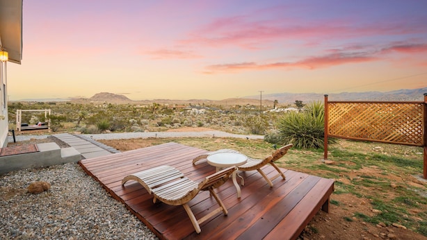 An outdoor patio with a wooden table and chairs, a small round table, and a view of a desert landscape with mountains in the distance. - Cholla Hills Home: Stunning 360° Views, Near JT (Yucca Valley)