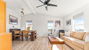 A spacious living room with a wooden floor, a beige sofa, a dining area with wooden chairs, a ceiling fan, and framed pictures on the walls. - Cholla Hills Home: Stunning 360° Views, Near JT (Yucca Valley)