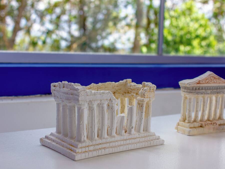 A white architectural model of ancient columns and a temple base with "Athens" inscribed on it, placed on a white surface near a window with a blue frame.