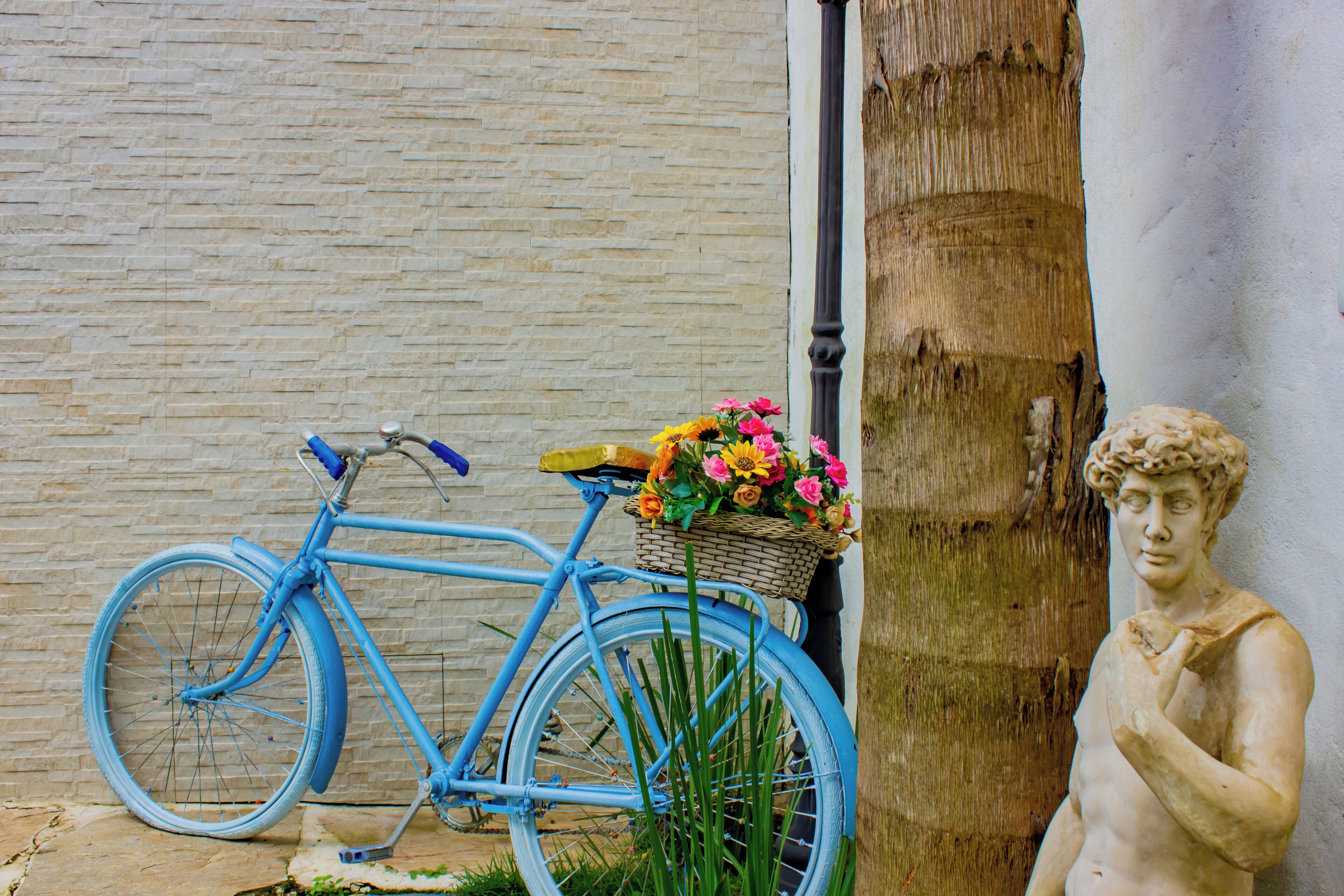 A blue bicycle with a basket of flowers, a statue, and a brick wall.