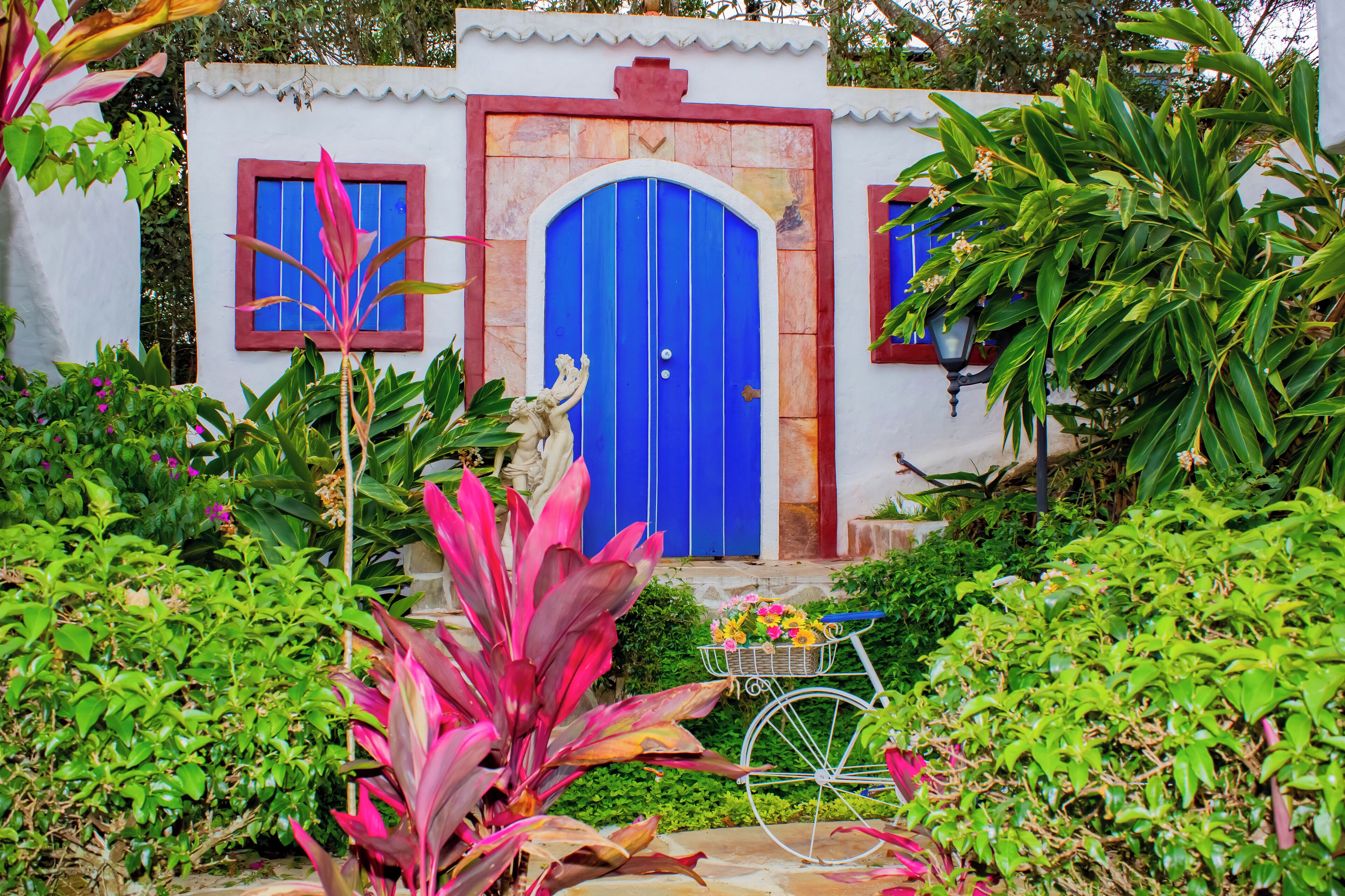A house with a blue door, surrounded by lush greenery and a bicycle with a basket of flowers.