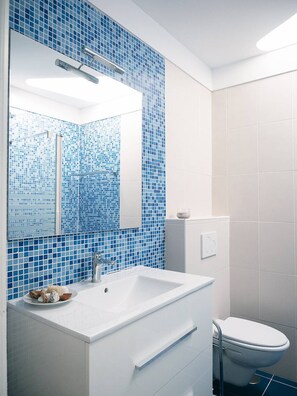 Ein Badezimmer mit einem weißen Waschbecken, einem Spiegel und einer Toilette. Die Wände sind mit blauen Mosaikfliesen versehen.