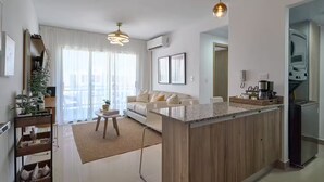 Un appartement moderne comprenant une cuisine, un coin repas et un salon. Il est doté d’un îlot central, d’une machine à café et d’une grande fenêtre avec des rideaux.