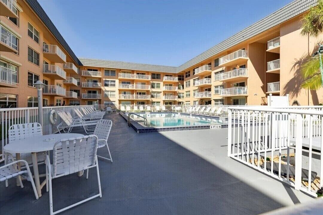 Un espace piscine avec des chaises et des tables blanches, entouré d’un immeuble à plusieurs étages doté de balcons.
