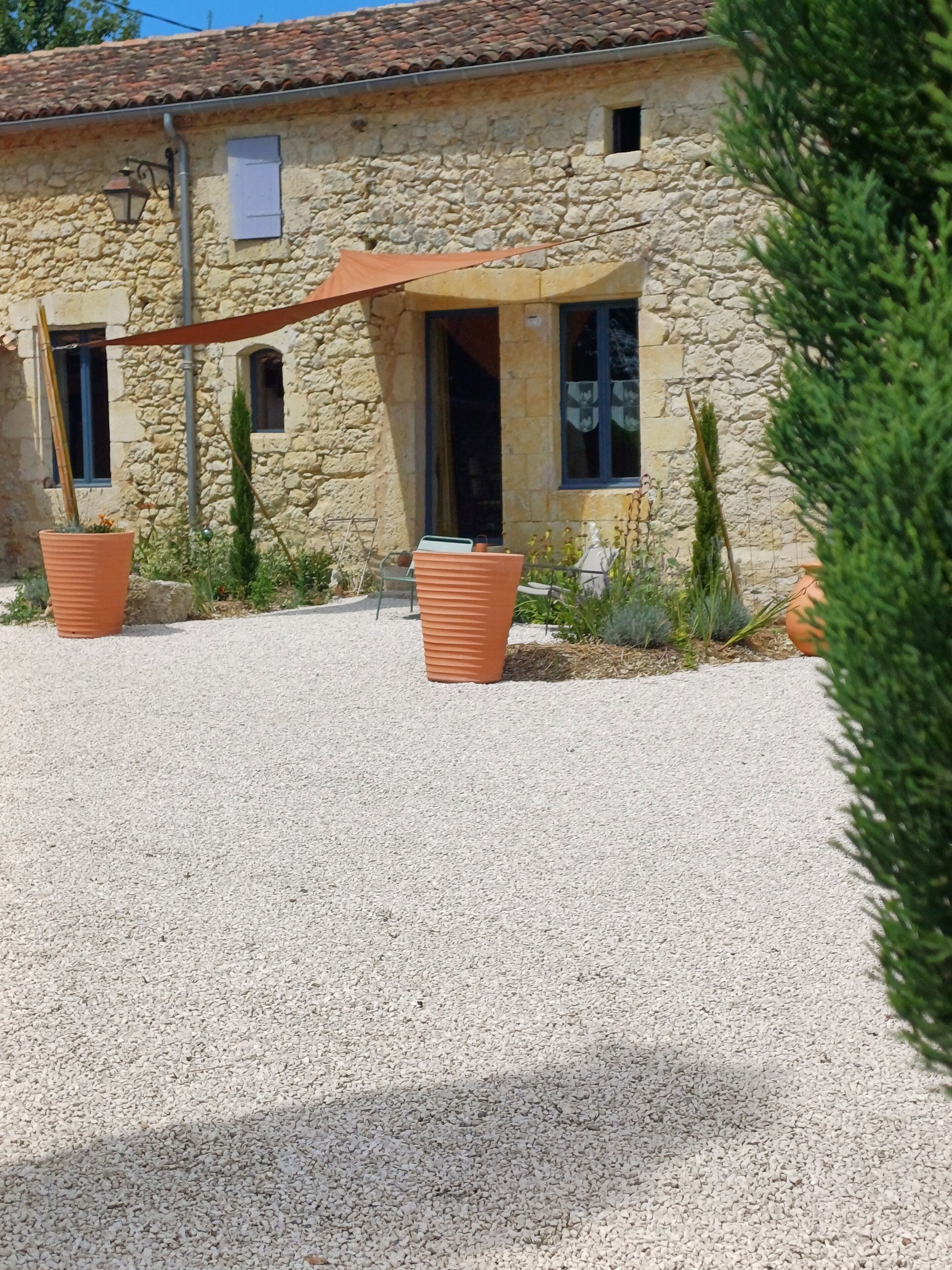 Een stenen gebouw met een grindpad, twee grote terracotta plantenbakken en een houten pergola.