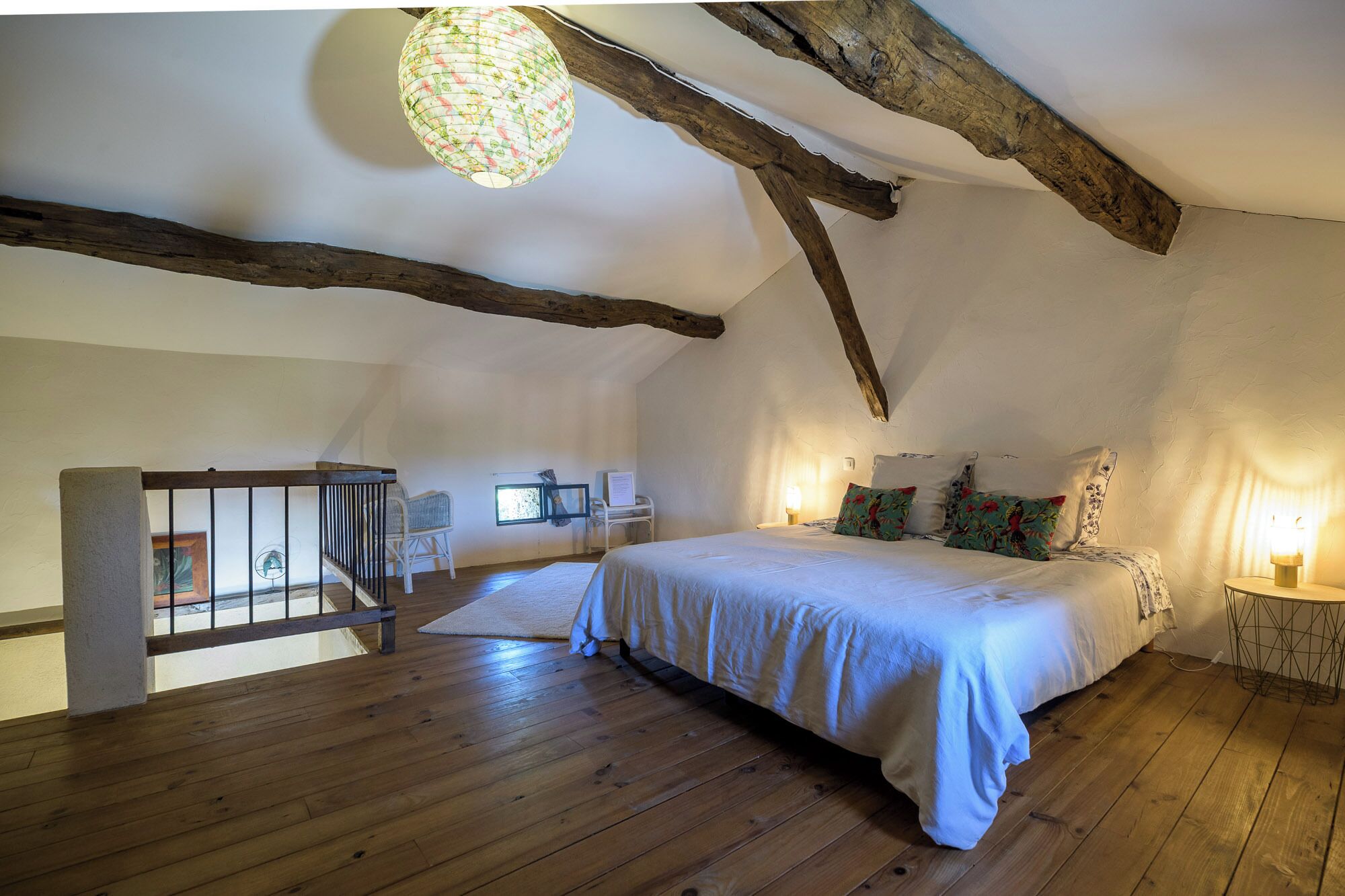 A bedroom with a wooden floor, a bed with white bedding, a hanging lantern, and exposed wooden beams.