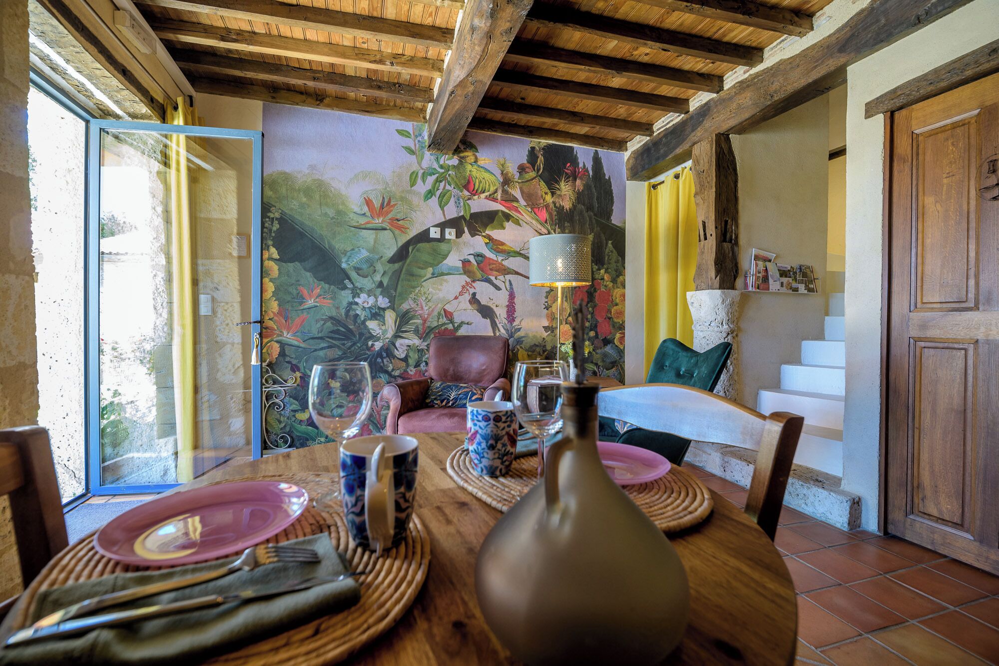 Un coin repas avec une table en bois dressée pour deux personnes, une fresque représentant des oiseaux et des fleurs sur le mur, et un escalier menant à un niveau supérieur.
