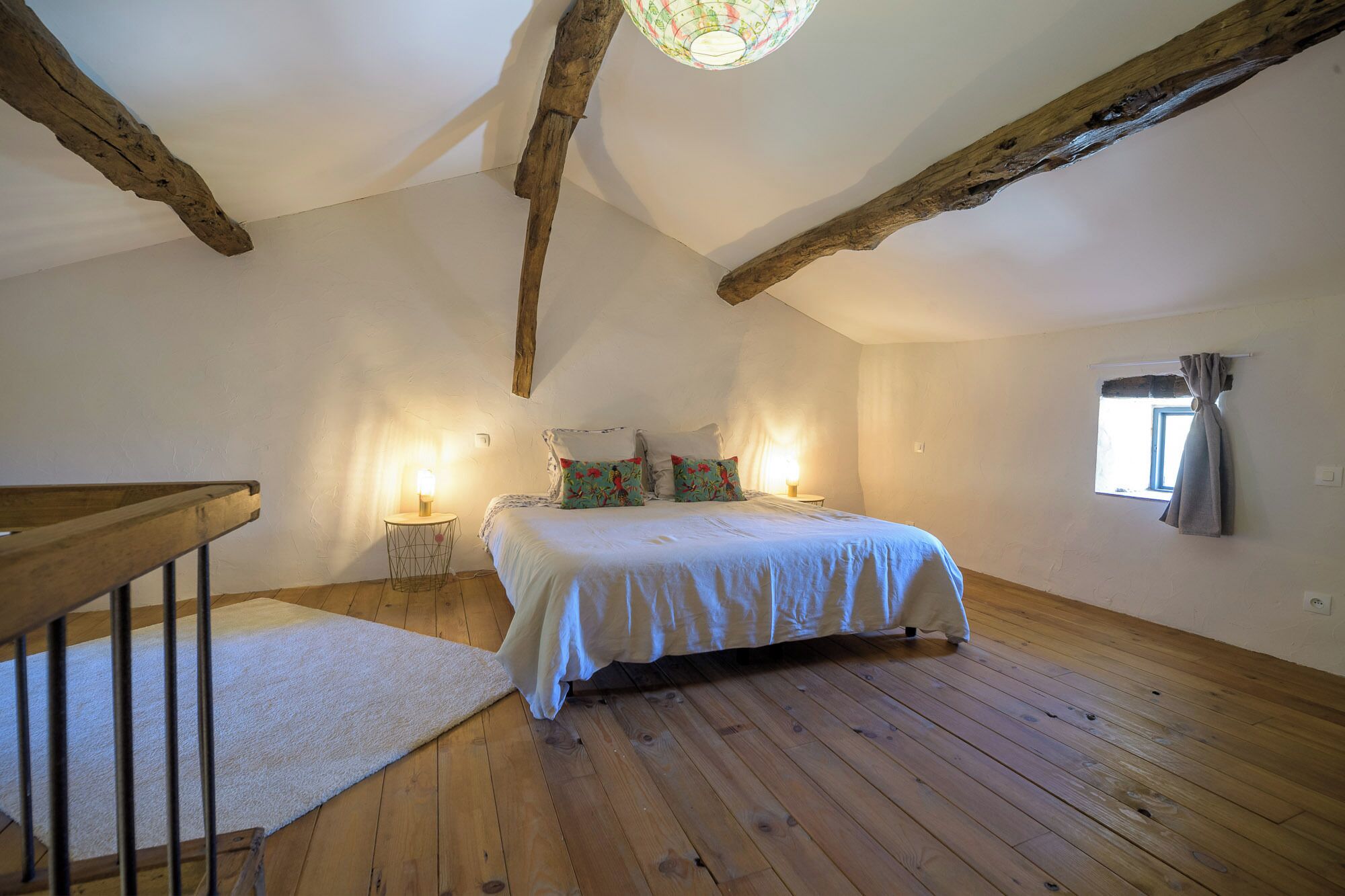 Un dormitorio con piso de madera, una cama con ropa blanca, dos lámparas de noche y una ventana con cortinas.